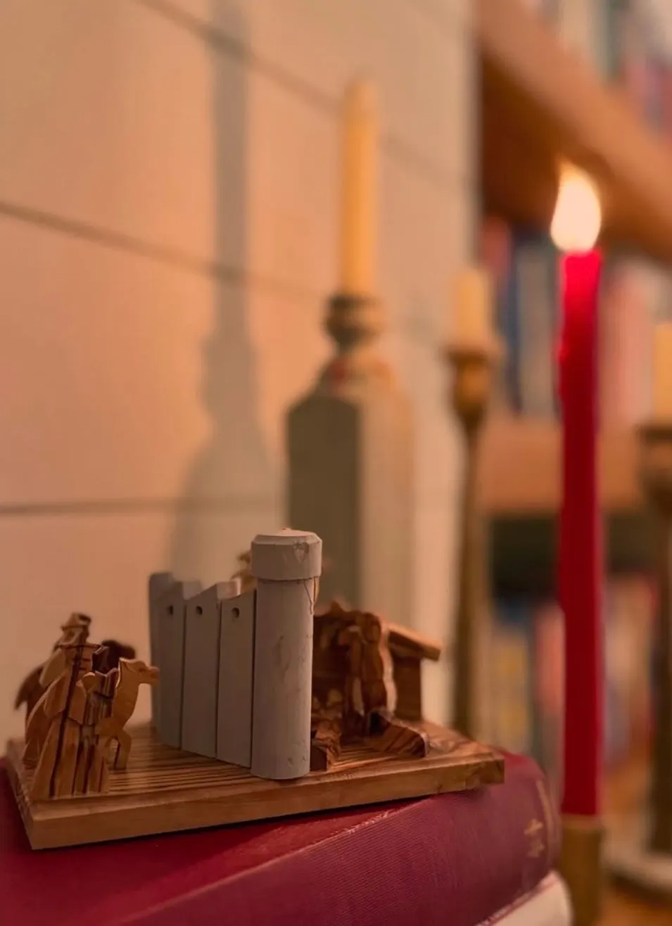 A red candle burning beside an olive wood nativity on a bookshelf